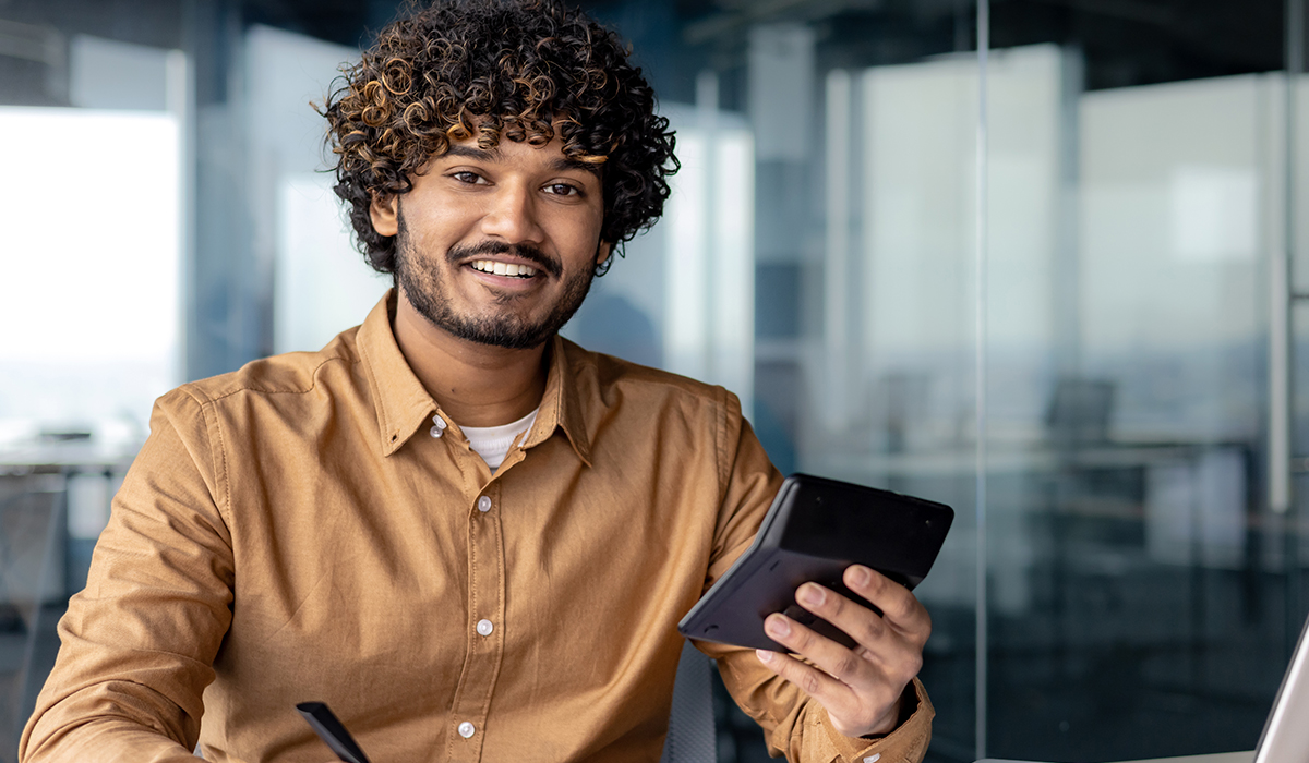 A smiling man holds a tablet in a modern office setting, with glass walls visible in the background.