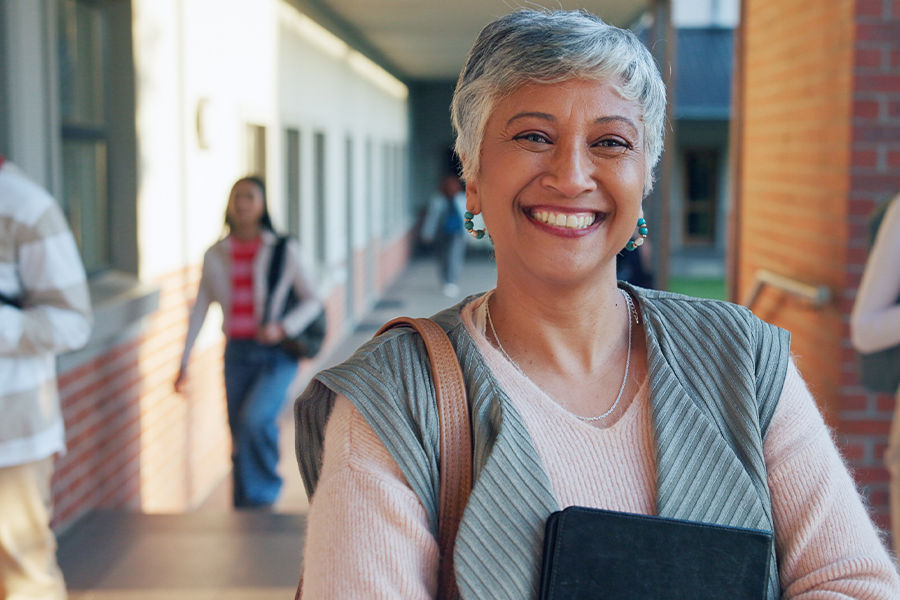 A smiling adult woman standing outdoors on a college campus walkway holding a notebook, with other students visible in the background.
