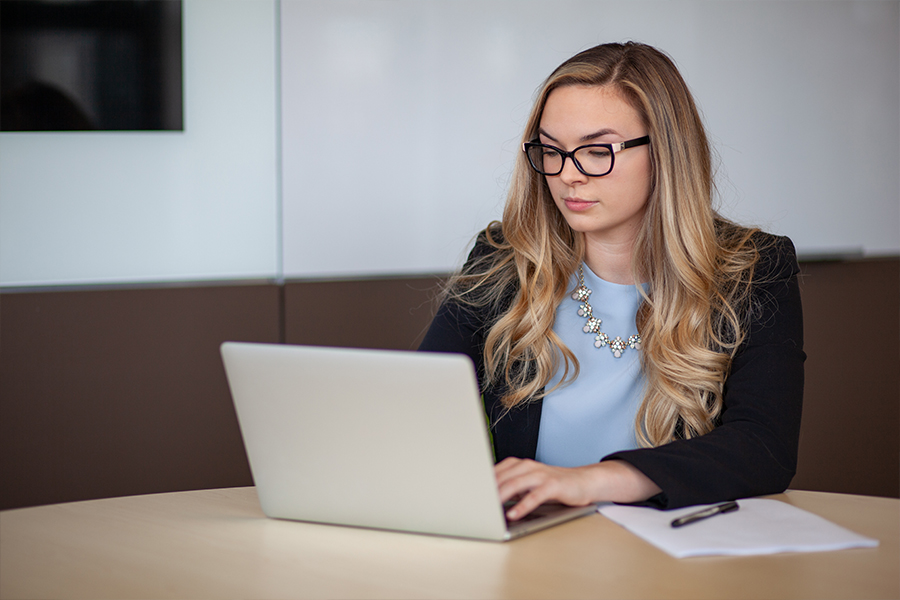 A woman in a blazer and glasses works on a laptop at a conference table with a notepad beside her.