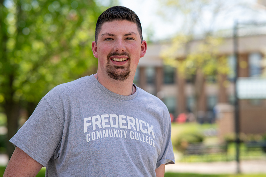 A smiling male student wearing a Frederick Community College t-shirt stands outdoors on campus on a sunny spring day.