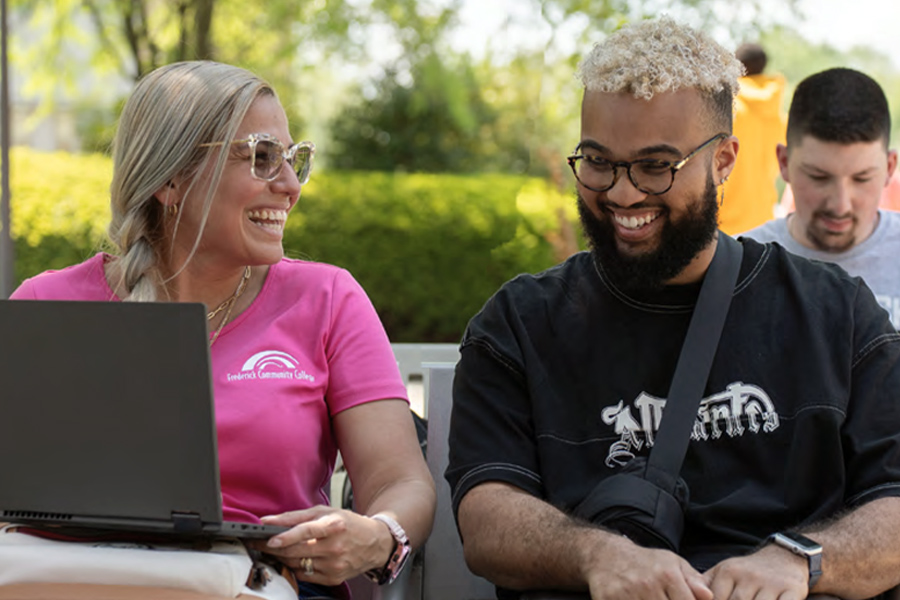 Two students laugh together while looking at a laptop outdoors on campus. One wears a pink Frederick Community College T-shirt.