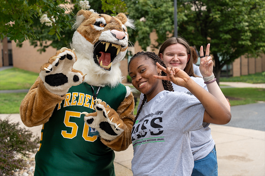 The Frederick Community College cougar mascot poses outdoors with two smiling students. The mascot, wearing a green and gold jersey with the number 50, playfully holds up its paws while the students stand beside it, both making peace signs. The student in the foreground wears a grey "PASS Program" t-shirt. Green trees and a campus building are visible in the background.