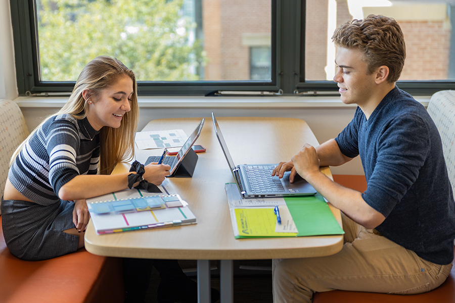 Four diverse students smile together outdoors in front of a campus building. They are wearing casual clothing, including Frederick Community College Cougars and Student Ambassador shirts.
