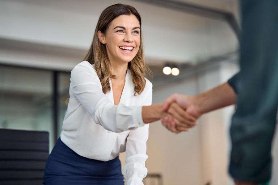 A smiling woman in a white blouse and navy skirt shaking hands with an unseen person in a professional office setting, conveying a successful meeting or greeting.