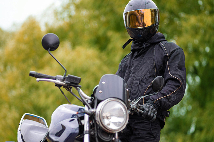 A motorcyclist in full black riding gear and a helmet with a gold-tinted visor stands beside a silver motorcycle with trees in the background.