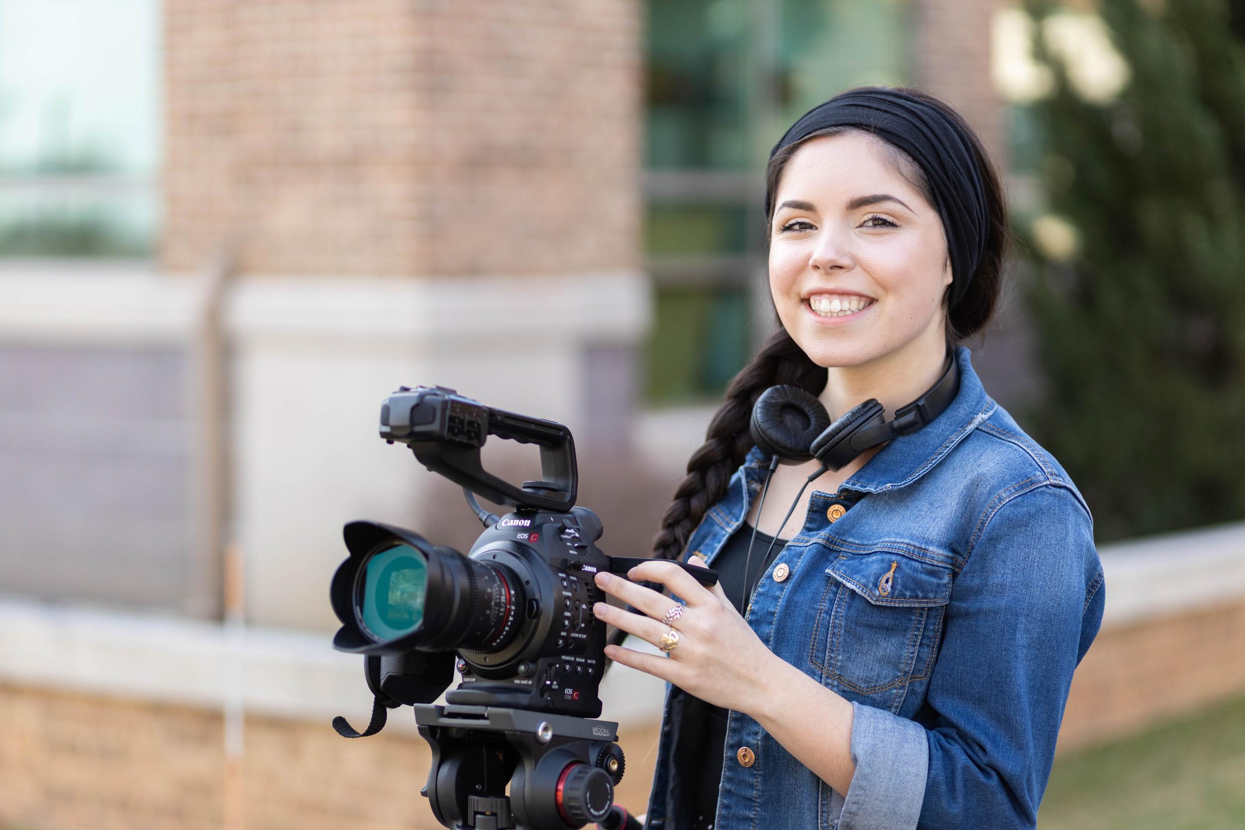 Woman in denim jacket, black scarf on head holding camera.